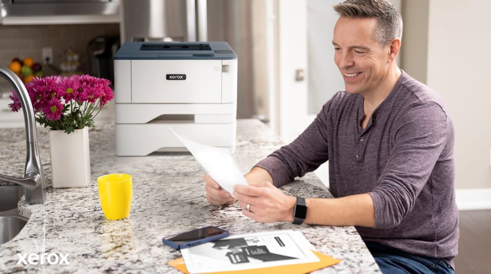Une personne sourit tout en examinant un document sur un comptoir de cuisine, avec une imprimante Xerox compacte et une tasse jaune vif à proximité, suggérant une impression et une gestion de documents transparentes dans un environnement domestique ou de petit bureau.